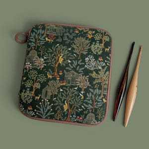 May include: A square zippered case featuring a dark green fabric with a repeating pattern of trees, greenhouses, birds, and small animals, accompanied by two crochet hooks, one dark brown and one light tan, positioned to the right of the case.