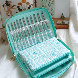 Puede incluir: Un estuche con cremallera de color verde menta que contiene un juego de agujas de tejer metálicas organizadas en ranuras individuales. El estuche cuenta con una solapa de tela blanca bordada con flores y etiquetas de 2mm a 5mm.