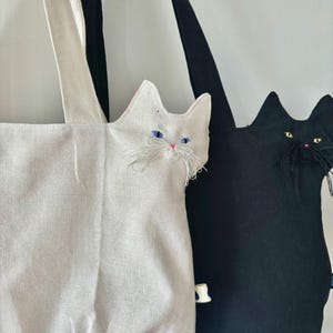 May include: Two cat-shaped tote bags. One is white with blue eyes and a pink nose, the other is black with yellow eyes and a pink nose. Both bags have long handles and are made of fabric.
