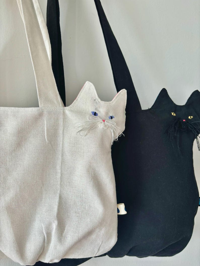 May include: Two cat-shaped tote bags. One is white with blue eyes and a pink nose, the other is black with yellow eyes and a pink nose. Both bags have long handles and are made of fabric.