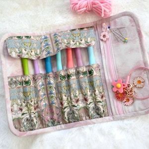 May include: Open crochet hook case with a floral pattern in pastel shades. The case displays colourful crochet hooks. A pink zip and flower-shaped zip pull are visible. Accessories include pins and daisy charms.