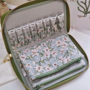 May include: Open green zippered case with multiple compartments. The interior features a floral pattern with pink and white lilies on a light blue background. The case has labeled pockets for knitting needles, and a zippered pouch.