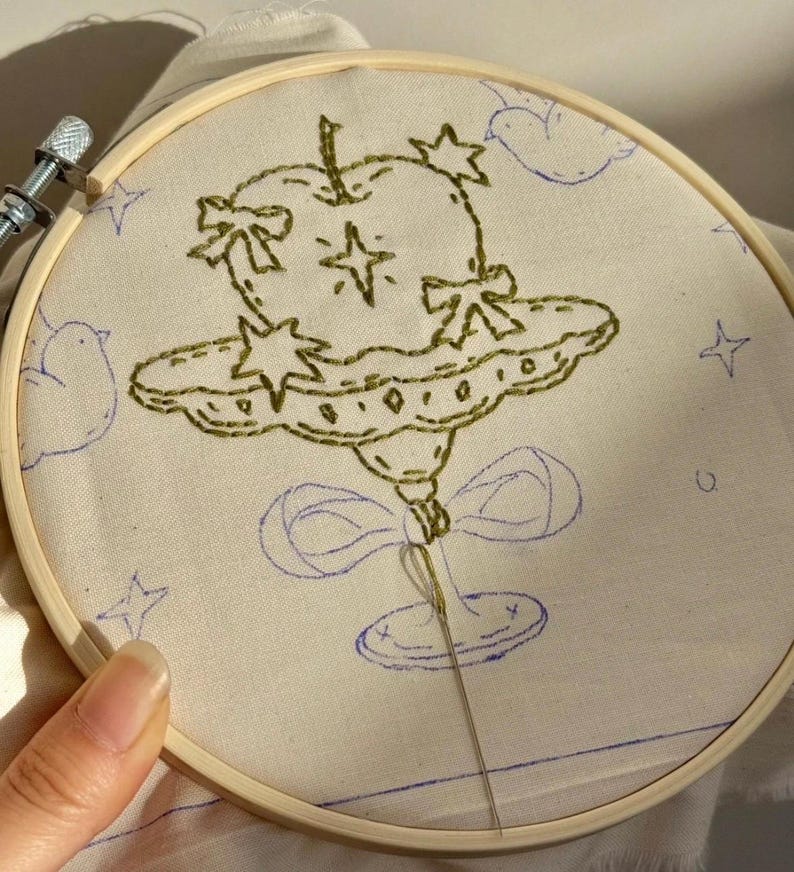 Pu&ograve; includere: Un telaio da ricamo con un design stravagante. Il design presenta un oggetto verde a forma di mela con stelle e fiocchi, che fluttua sopra una forma di nuvola. Contorni blu di nuvole e stelle circondano l'immagine centrale. Una mano tiene il telaio.