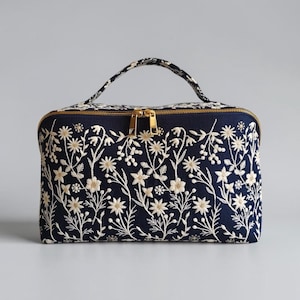 May include: A rectangular navy blue fabric bag featuring an all-over cream-coloured floral embroidery pattern. The bag includes a top handle, a gold-toned metal zip closure with two pulls, and a structured shape with rounded corners.