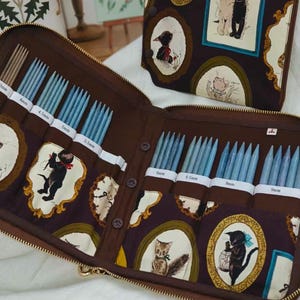 Puede incluir: Estuche marrón abierto con cremallera para agujas de tejer con agujas azules de varios tamaños. El estuche está decorado con ilustraciones de gatos enmarcadas. Un estuche a juego es visible en el fondo. Las agujas están etiquetadas con tamaños en milímetros.