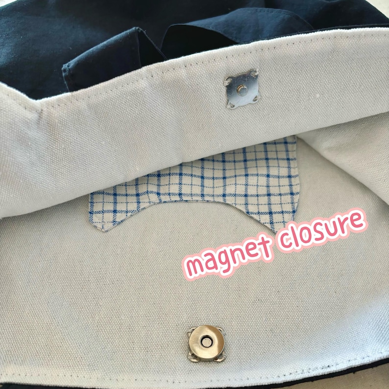 May include: Close-up of a navy blue tote bag interior with a light-colored lining. The bag features a magnetic closure. A small, blue and white plaid fabric detail is visible inside the bag.