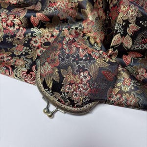 May include: A black fabric with a floral pattern in red and gold is displayed, with a metal clasp purse. The purse is open, revealing the same floral pattern on its lining. The fabric is folded, showing the intricate details of the design.