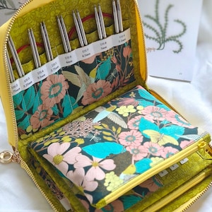 Peut inclure: Un étui jaune à fermeture éclair contenant un ensemble d'aiguilles à tricoter circulaires en métal. L'intérieur présente un tissu sombre à motif floral et colibri, avec des emplacements étiquetés pour les tailles US 2 à US 9, de 2,75mm à 5,5mm.