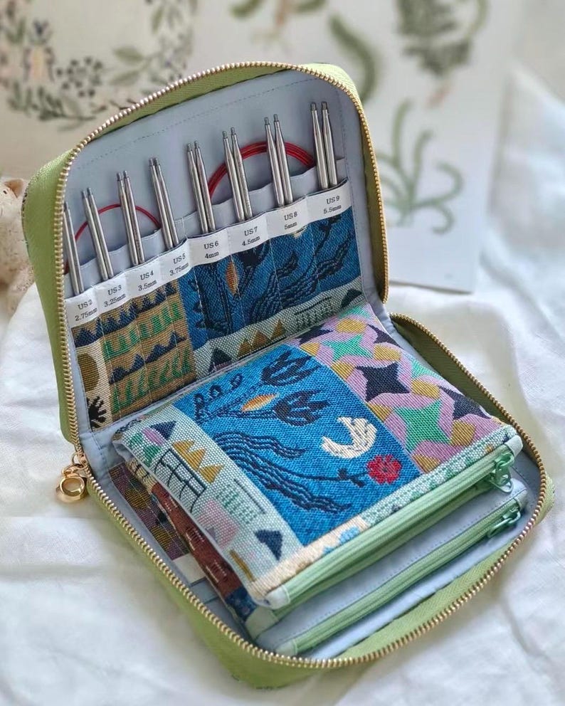 K&ouml;nnte beinhalten: Ein salbeigr&uuml;nes Etui mit Rei&szlig;verschluss enth&auml;lt ein Set austauschbarer Stricknadeln mit roten Kabeln. Das Innere bietet beschriftete F&auml;cher f&uuml;r die Gr&ouml;&szlig;en US 2 (2,75 mm) bis US 9 (5,5 mm) sowie bunte Stofftaschen mit floralen und geometrischen Mustern.