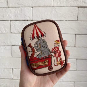 Könnte beinhalten: Eine beige, rechteckige Geldbörse mit einer Illustration zum Thema Zirkus. Das Design zeigt einen Elefanten, einen Clown und einen Affen auf einem roten Wagen unter einem rot-weiß gestreiften Regenschirm. Die Geldbörse hat einen braunen Rand.