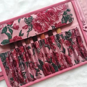 May include: A pink, floral-patterned roll-up case, possibly for art supplies. The case has a repeating pattern of red roses and green leaves on a pink background. The case is open, revealing several coloured pencils or crayons.