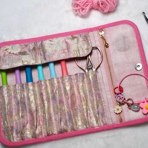 May include: A pink and gold fabric crochet hook case with a zipper closure, holding several colorful crochet hooks. The case also contains a pair of decorative scissors and a charm bracelet. A ball of pink yarn is in the background.
