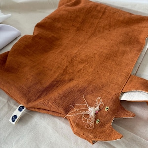 Op de afbeelding: Een oranje katvormig kussen met geborduurde details. Het kussen heeft een textuurstof en een witte binnenkant. Het gezicht van de kat heeft geborduurde ogen, snorharen en een roze neus. Een klein label met een blauw ontwerp is bevestigd.