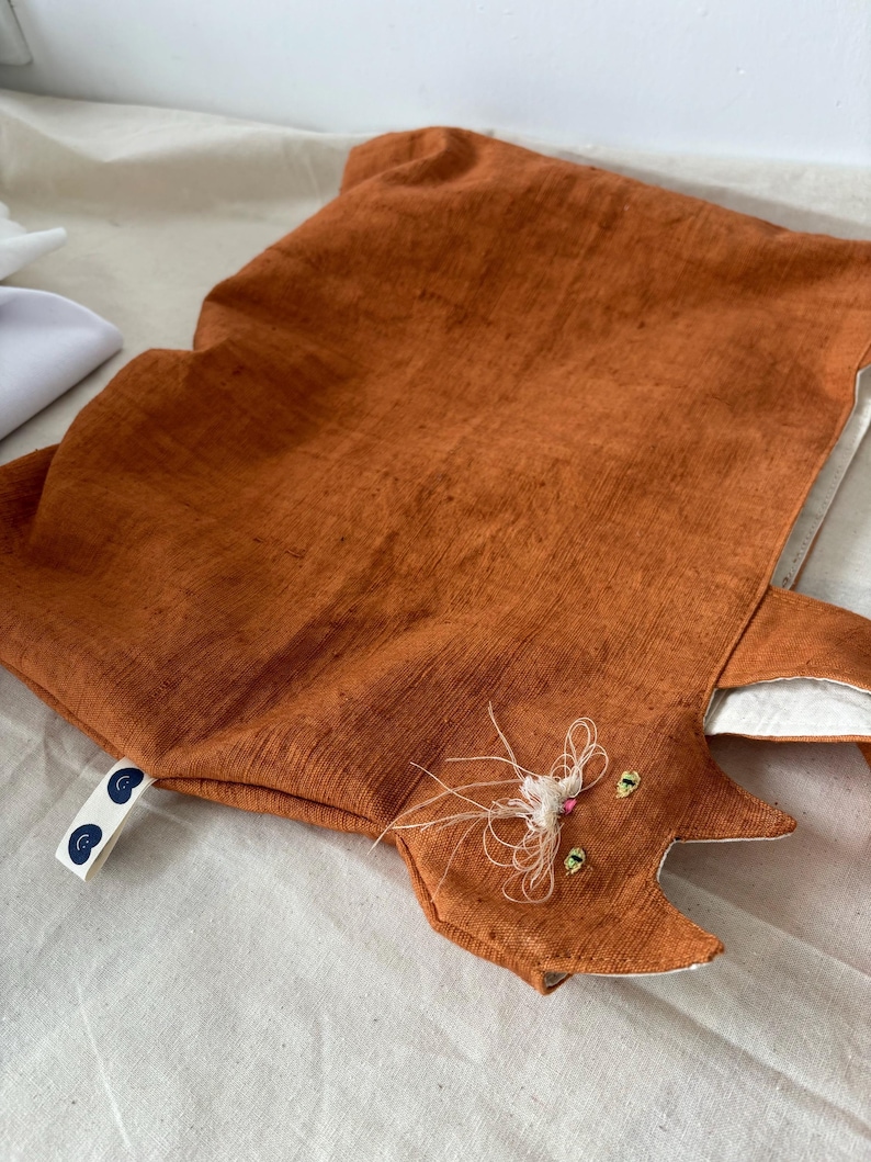 Op de afbeelding: Een oranje katvormig kussen met geborduurde details. Het kussen heeft een textuurstof en een witte binnenkant. Het gezicht van de kat heeft geborduurde ogen, snorharen en een roze neus. Een klein label met een blauw ontwerp is bevestigd.