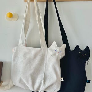 May include: Two cat-shaped tote bags hanging on wooden pegs. One bag is white with a white cat face and tail, the other is black with a black cat face and tail. Both bags have long straps for carrying.