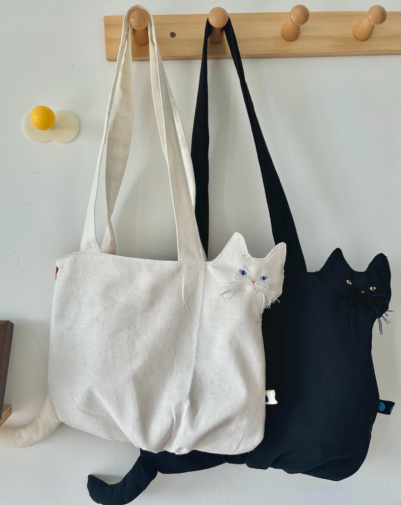 May include: Two cat-shaped tote bags hanging on wooden pegs. One bag is white with a white cat face and tail, the other is black with a black cat face and tail. Both bags have long straps for carrying.