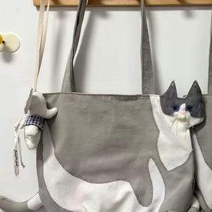 May include: Gray and white cat-shaped tote bag with long handles. The bag features a plush cat face and tail, with a small stuffed animal hanging from the handle. The bag is made of a soft fabric.
