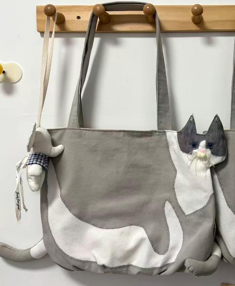 May include: Gray and white cat-shaped tote bag with long handles. The bag features a plush cat face and tail, with a small stuffed animal hanging from the handle. The bag is made of a soft fabric.