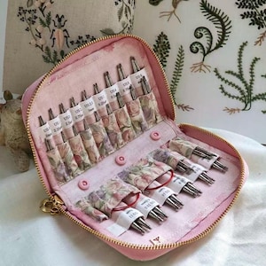 May include: A pink zippered case containing a set of interchangeable knitting needles. The interior features floral fabric pockets labeled with US sizes ranging from 2mm to 11, holding metal needle tips and red circular cables for knitting projects.