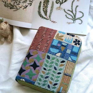K&ouml;nnte beinhalten: Ein rechteckiges Etui mit Rei&szlig;verschluss und einem Patchwork-Design aus gewebten Stoffbahnen. Die Muster umfassen geometrische Formen, stilisierte Bl&auml;tter und abstrakte blaue Motive in Braun-, Gr&uuml;n-, Lila- und Blaut&ouml;nen mit gr&uuml;nem Rand.
