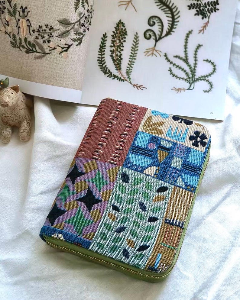 K&ouml;nnte beinhalten: Ein rechteckiges Etui mit Rei&szlig;verschluss und einem Patchwork-Design aus gewebten Stoffbahnen. Die Muster umfassen geometrische Formen, stilisierte Bl&auml;tter und abstrakte blaue Motive in Braun-, Gr&uuml;n-, Lila- und Blaut&ouml;nen mit gr&uuml;nem Rand.