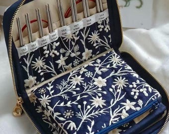 Puede incluir: Un estuche azul marino con cremallera y bordado floral blanco, que contiene un juego de agujas de tejer circulares de metal. Las ranuras interiores están etiquetadas con tallas de US 2 (2,75 mm) a US 9 (5,5 mm), con cables rojos.