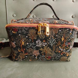 May include: A black and gold patterned handbag with a top handle and a detachable strap. The bag features a forest scene with trees, birds, and small buildings. The zipper is gold-colored.