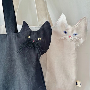 May include: Two tote bags with cat-shaped appliqu&eacute;s. One bag is black with yellow eyes and a black mustache, the other is off-white with blue eyes and white whiskers. Both bags have long straps and a small tag with a blue and white polka dot pattern.
