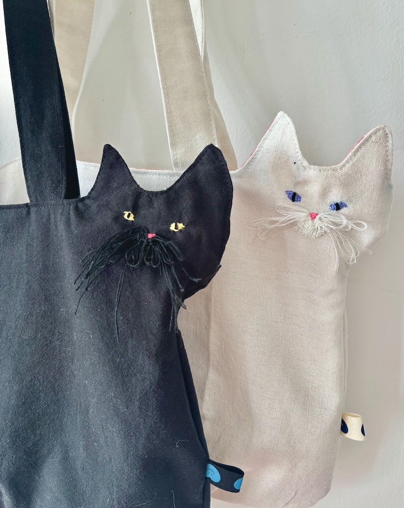 May include: Two tote bags with cat-shaped appliqu&eacute;s. One bag is black with yellow eyes and a black mustache, the other is off-white with blue eyes and white whiskers. Both bags have long straps and a small tag with a blue and white polka dot pattern.