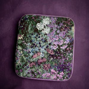 Pode incluir: Um estojo quadrado com fecho de correr apresentando uma estampa floral pictórica vibrante em tons de roxo, rosa, branco e verde. O design retrata uma cena de jardim densa com várias pequenas flores e folhagens, com acabamento em tecido roxo claro.