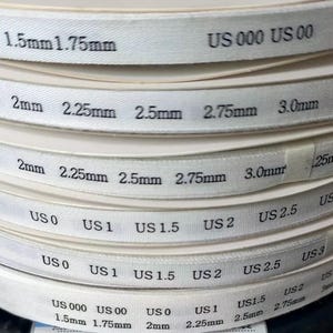 K&ouml;nnte beinhalten: Ein Stapel wei&szlig;er Ma&szlig;b&auml;nder f&uuml;r Stricknadeln mit schwarzem Aufdruck, der die Gr&ouml;&szlig;en in Millimetern von 1,5 mm bis 3,25 mm und US-Gr&ouml;&szlig;en von US 000 bis US 4 angibt. Die B&auml;nder sind in Schichten aufgerollt und zeigen deutlich lesbare Zahlen.