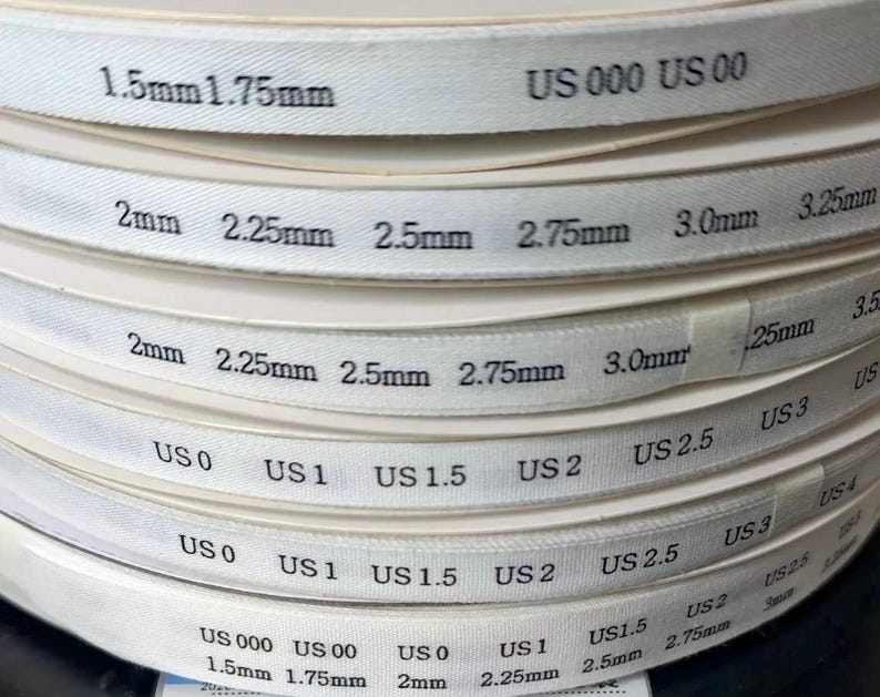 K&ouml;nnte beinhalten: Ein Stapel wei&szlig;er Ma&szlig;b&auml;nder f&uuml;r Stricknadeln mit schwarzem Aufdruck, der die Gr&ouml;&szlig;en in Millimetern von 1,5 mm bis 3,25 mm und US-Gr&ouml;&szlig;en von US 000 bis US 4 angibt. Die B&auml;nder sind in Schichten aufgerollt und zeigen deutlich lesbare Zahlen.
