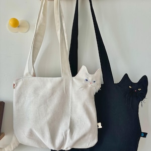 May include: Two cat-shaped tote bags hanging on a wooden rack. One bag is off-white, the other is black. Both bags have long straps and cat features, including ears, eyes, and a tail. The off-white bag has blue eyes, and the black bag has yellow eyes.