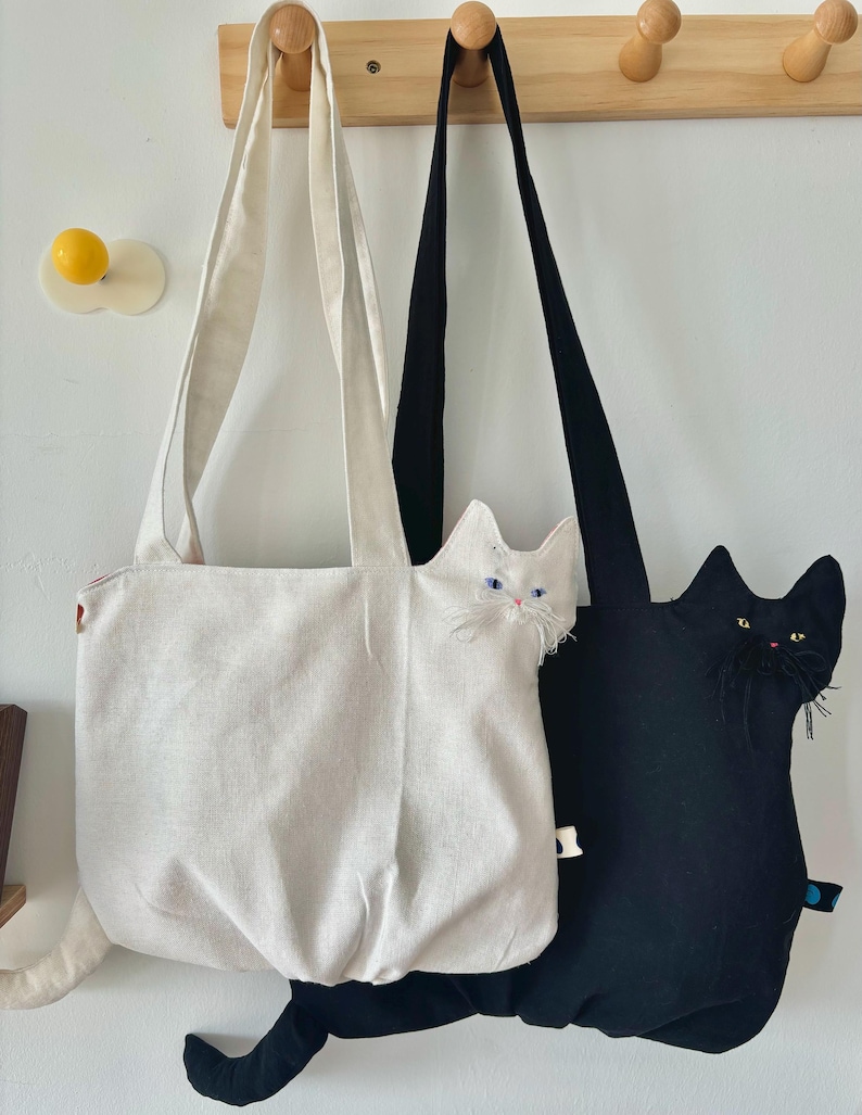 May include: Two cat-shaped tote bags hanging on a wooden rack. One bag is off-white, the other is black. Both bags have long straps and cat features, including ears, eyes, and a tail. The off-white bag has blue eyes, and the black bag has yellow eyes.