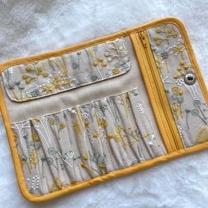 May include: A beige pencil case with embroidered floral detailing in yellow, white, and green. It has a yellow zip and trim, multiple pockets, and a snap closure. The case is open, showing the interior. The case is approximately 25 cm long.