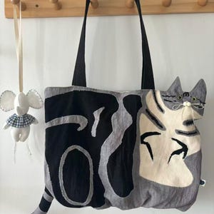 May include: A gray and black tote bag with a cat design. The bag features a large cat face on one side and a tail extending from the bottom. The bag has black handles and is hanging on a wooden rack. A small stuffed elephant is hanging next to the bag.