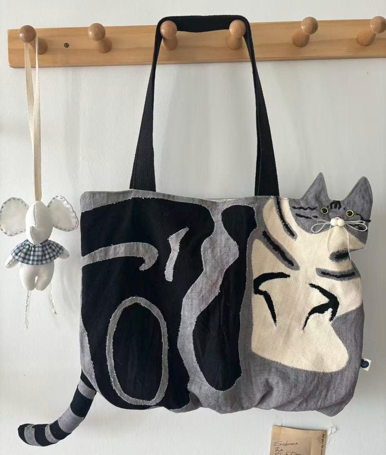 May include: A gray and black tote bag with a cat design. The bag features a large cat face on one side and a tail extending from the bottom. The bag has black handles and is hanging on a wooden rack. A small stuffed elephant is hanging next to the bag.