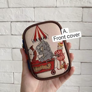 Puede incluir: Una cubierta frontal rectangular con un diseño de circo. La cubierta presenta una ilustración de un elefante, un payaso y un vagón de circo rojo bajo una carpa a rayas rojas y blancas. El texto "A. Front cover" es visible.