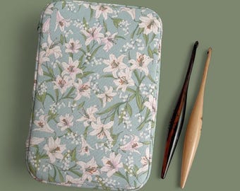 May include: A light blue floral case with a zipper, featuring a pattern of white and pink lilies and green leaves. Two wooden crochet hooks, one dark brown and one light brown, are next to the case. The background is a solid olive green.