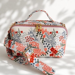 Puede incluir: Un bolso bandolera rectangular con base de color crema y bordados florales intrincados en tonos rojo, coral y gris. El bolso incluye un asa superior, una correa de hombro ancha ajustable y un cierre de cremallera en tono dorado.