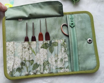 Puede incluir: Un estuche verde de tela para ganchillos con un bolsillo con estampado floral y cuatro ganchillos. El estuche tiene cierre de cremallera, cierre de botón a presión y un par de tijeras. El estuche está abierto, revelando el contenido.