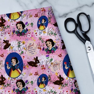 Puede incluir: Tela rosa con un patrón repetido de Blancanieves, ciervos y elementos florales. El diseño incluye el texto "Do good and good will come to you". Tijeras negras están situadas en el lado derecho de la imagen.