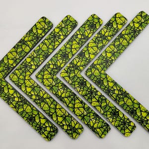 May include: A collection of eight L-shaped wooden coasters. Each coaster features a vibrant green and yellow leaf pattern with black vein-like lines. The coasters are arranged in a chevron pattern against a white background.