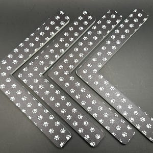 May include: Six black L-shaped corner protectors with a repeating pattern of white paw prints. The protectors are arranged in a diagonal pattern against a dark background, suggesting a pet-themed product.
