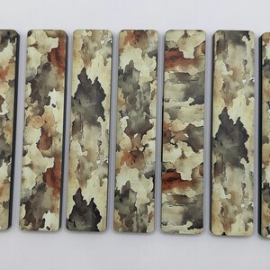 May include: Nine rectangular decorative wooden pieces with a camouflage pattern in shades of brown, beige, and gray. The pieces are arranged horizontally against a white background, showcasing their unique design.