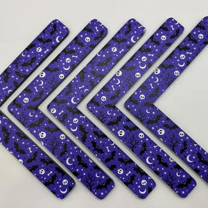 May include: Six purple, L-shaped Halloween-themed decorations. Each piece features a repeating pattern of black bats, skulls, bones, moons, stars, and trees. The decorations are arranged in a chevron pattern against a white background.