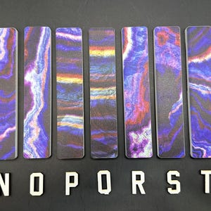 May include: Nine rectangular bookmarks with a marbled design in shades of purple, red, and blue. The bookmarks are arranged vertically on a black surface, with white letters spelling out "MNOPRSTU" below them. The bookmarks have a unique, artistic appearance.