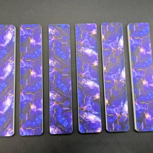 May include: Six rectangular bookmarks with a marbled design in shades of purple, blue, and gold. The bookmarks have a white border and a smooth finish. The design resembles a galaxy or nebula.