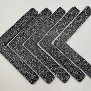 May include: A collection of seven black and white decorative corner pieces. Each piece is shaped like an L, with a pattern of white spiderwebs on a black background. The pieces are arranged in a diagonal line, creating a visually appealing display.