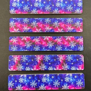 May include: Five rectangular name tags with a vibrant design. The background features a blend of blue and pink hues, resembling a galaxy. White snowflakes are scattered across the surface, creating a wintery theme. The name tags are arranged vertically against a dark background.